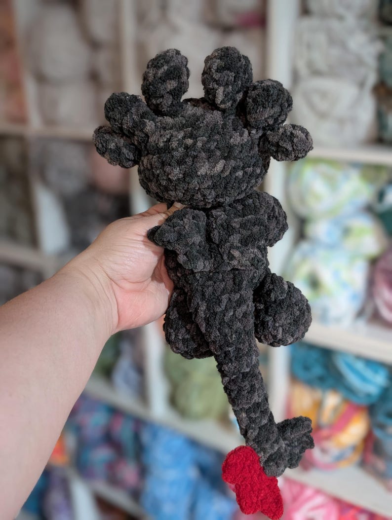 May include: A handmade, dark gray crocheted dragon plushie with a red tail. The dragon has a textured, plush appearance, with a head featuring multiple rounded protrusions. The tail is a vibrant red, contrasting with the gray body. The plushie is held in a hand.