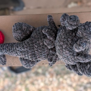 May include: A handmade crochet dragon toy in shades of gray with a bright red tail. The dragon has a textured, plush appearance, with detailed wings and limbs. The toy is lying on a wooden surface.