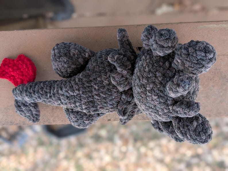May include: A handmade crochet dragon toy in shades of gray with a bright red tail. The dragon has a textured, plush appearance, with detailed wings and limbs. The toy is lying on a wooden surface.