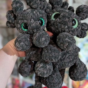 May include: Two handmade, dark gray plush dragon toys with green glitter eyes and small red accents. The plush toys have a soft, textured appearance and are held up in front of a blurred background.