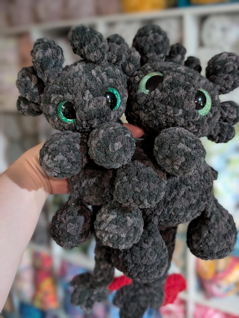 May include: Two handmade, dark gray plush dragon toys with green glitter eyes and small red accents. The plush toys have a soft, textured appearance and are held up in front of a blurred background.