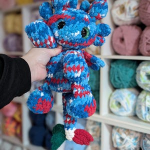 May include: A handmade plush creature in blue, red, and white yarn. The toy has large, round eyes and a whimsical design. The plushie is held up in front of a shelf filled with yarn.
