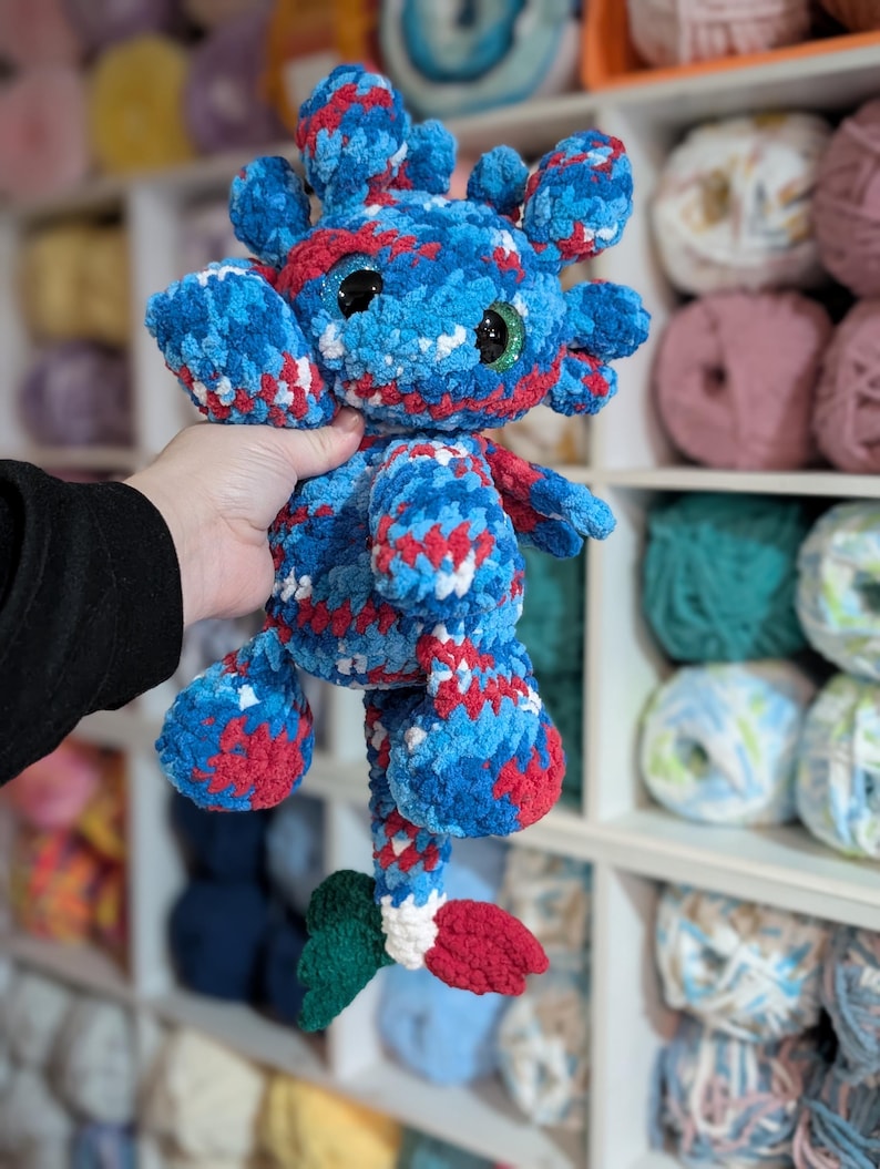 May include: A handmade plush creature in blue, red, and white yarn. The toy has large, round eyes and a whimsical design. The plushie is held up in front of a shelf filled with yarn.