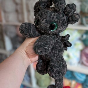 May include: A handmade, plush dragon toy in shades of gray and black. The dragon has large, glittery green eyes, small wings, and a red tail tip. The toy is made of a soft, textured yarn, and is held in a person's hand.
