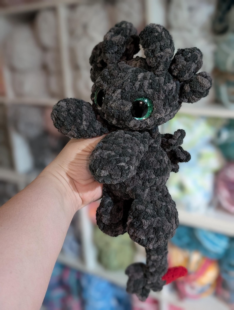 May include: A handmade, plush dragon toy in shades of gray and black. The dragon has large, glittery green eyes, small wings, and a red tail tip. The toy is made of a soft, textured yarn, and is held in a person's hand.