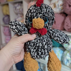 May include: A handmade crochet chicken plushie with a black and white body, red comb and wattle, and orange beak and feet. The chicken has blue eyes and is made of soft yarn. The background shows shelves filled with yarn.