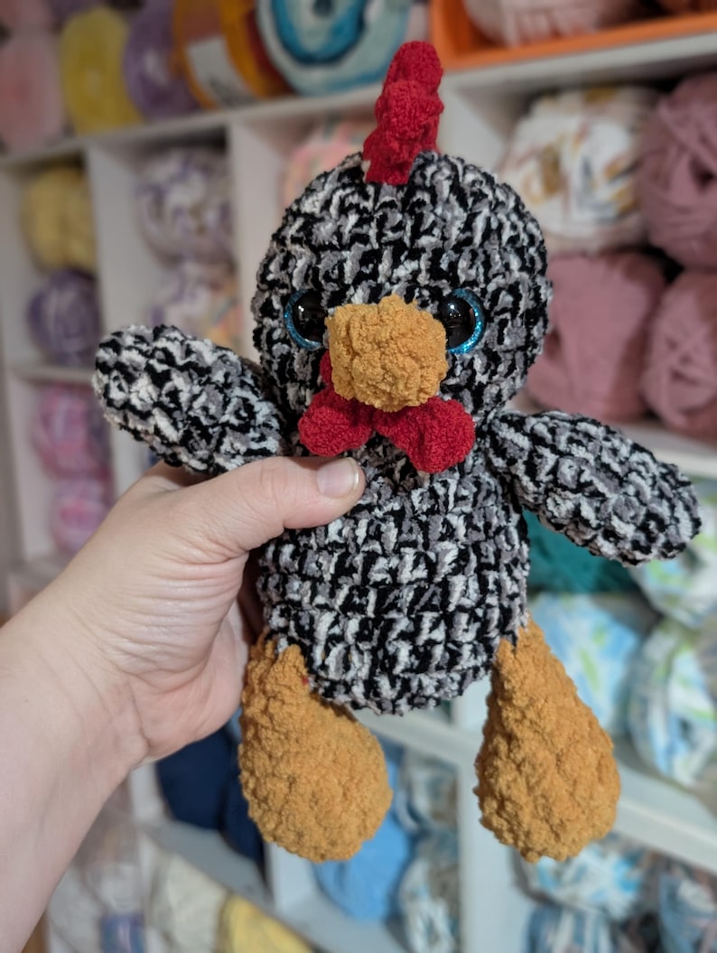 May include: A handmade crochet chicken plushie with a black and white body, red comb and wattle, and orange beak and feet. The chicken has blue eyes and is made of soft yarn. The background shows shelves filled with yarn.