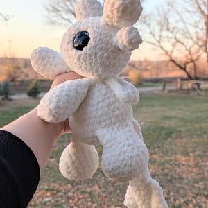 May include: A handmade white crochet plush toy with large black eyes and rounded features. The plush has a soft, textured appearance and is held up against a blurred outdoor background. The toy has a whimsical, cartoonish design.