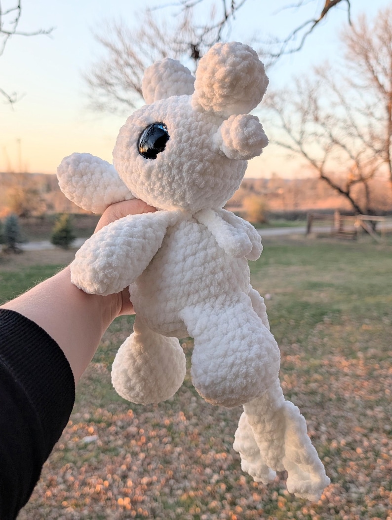 May include: A handmade white crochet plush toy with large black eyes and rounded features. The plush has a soft, textured appearance and is held up against a blurred outdoor background. The toy has a whimsical, cartoonish design.
