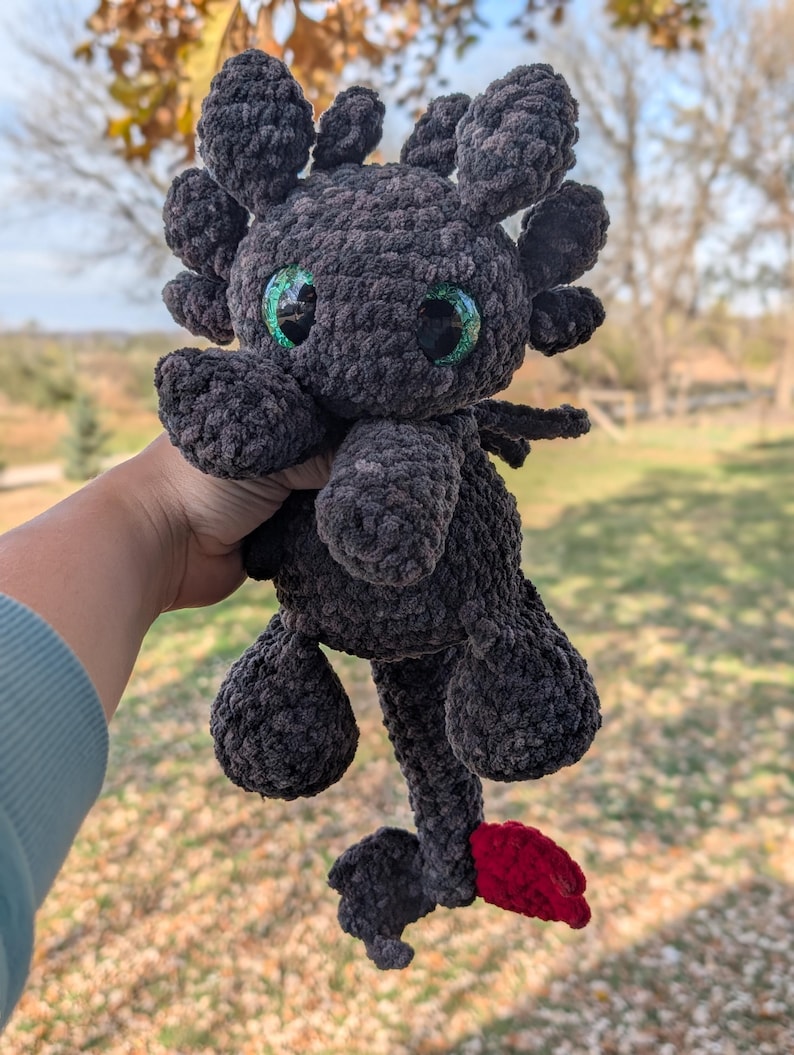 May include: A handmade, dark gray crochet dragon plushie with large, glittery green eyes and a red tail fin. The dragon has small wings and is held up against a blurred outdoor background.