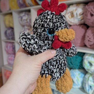May include: A handmade crochet chicken with a black, white, and gray body. It has a red comb and wattle, a yellow beak, and brown feet. The chicken has blue eyes. The background shows shelves filled with yarn.