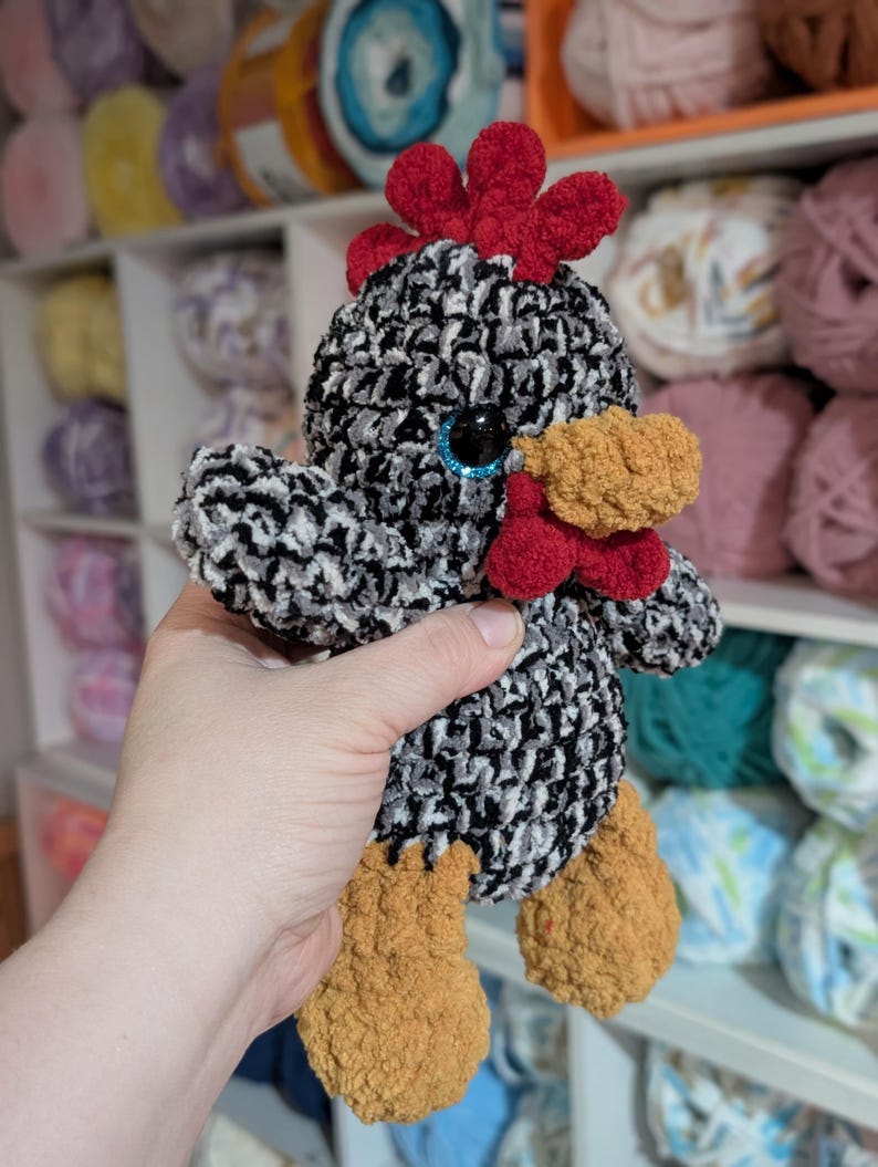 May include: A handmade crochet chicken with a black, white, and gray body. It has a red comb and wattle, a yellow beak, and brown feet. The chicken has blue eyes. The background shows shelves filled with yarn.