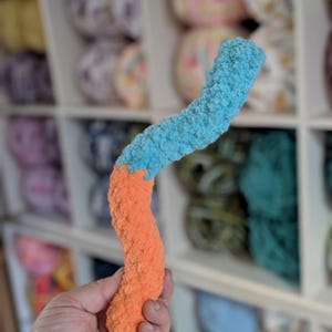 Puede incluir: Un juguete de gusano de peluche hecho a mano con una combinación de colores vibrantes. El gusano es principalmente naranja, que pasa a azul claro en la parte superior. El juguete tiene una apariencia texturizada y difusa, lo que sugiere una sensación suave y acogedora. El fondo muestra estantes llenos de hilo colorido.