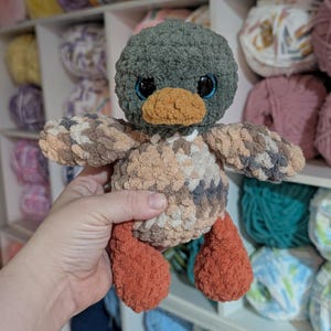 May include: A plush crochet duck toy featuring a dark green head, a tan beak, and large black eyes. The body is crafted from variegated yarn in shades of beige, brown, and gray, while the feet are a solid, textured burnt orange color.