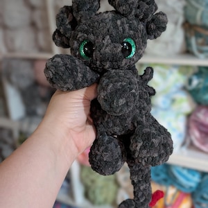 May include: A handmade, plush dragon toy in shades of gray and black. The dragon has large, glittery green eyes, small horns, and a long tail with a red tip. The toy is made of a soft, textured material, and is held in a person's hand.