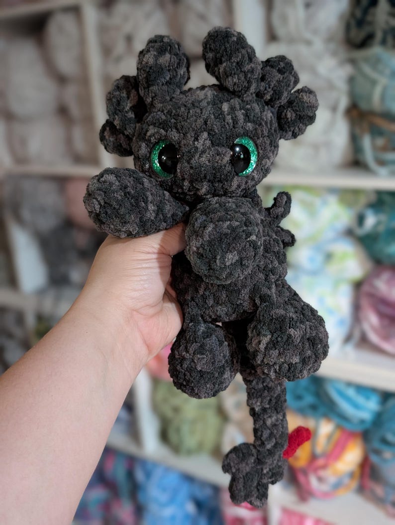 May include: A handmade, plush dragon toy in shades of gray and black. The dragon has large, glittery green eyes, small horns, and a long tail with a red tip. The toy is made of a soft, textured material, and is held in a person's hand.