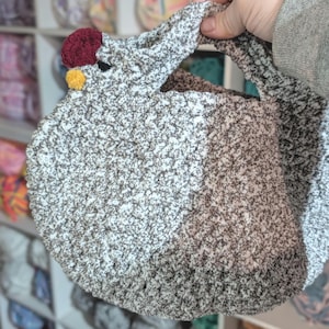 Puede incluir: Un bolso de ganchillo hecho a mano con forma de gallina. El bolso es principalmente blanco y marrón moteado, con una cresta roja y una carúncula amarilla. El asa está integrada en la cabeza de la gallina. El bolso lo sostiene una persona.