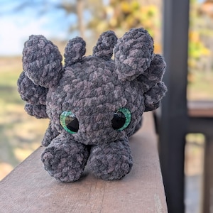 May include: A handmade plush axolotl in shades of gray, with large, expressive green eyes. The soft, textured material gives the creature a cuddly appearance. The axolotl is posed on a wooden surface, showcasing its unique design.