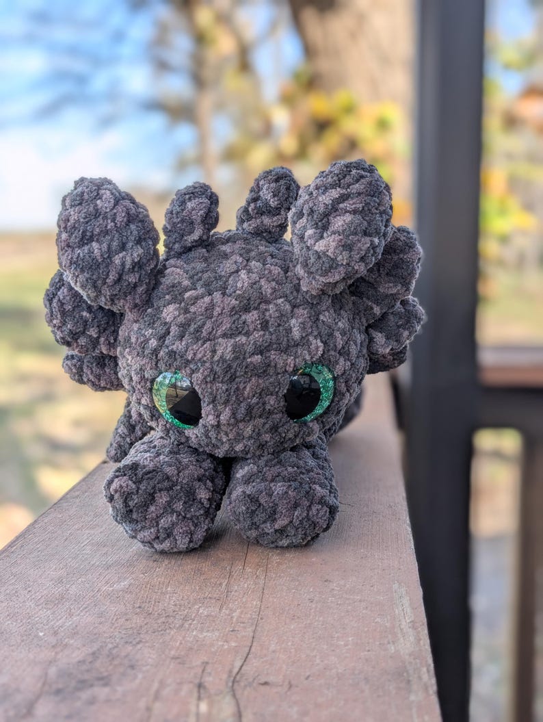 May include: A handmade plush axolotl in shades of gray, with large, expressive green eyes. The soft, textured material gives the creature a cuddly appearance. The axolotl is posed on a wooden surface, showcasing its unique design.