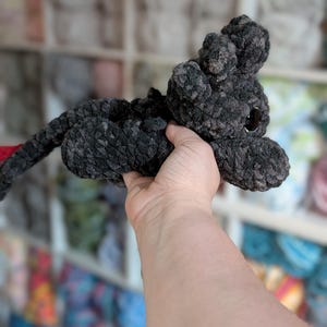 May include: A handmade, dark gray crocheted cat toy with a long tail and large, dark eyes. The cat is made of a soft, textured yarn and has small, rounded ears. The tail is red. The toy is held up in front of a blurred background.
