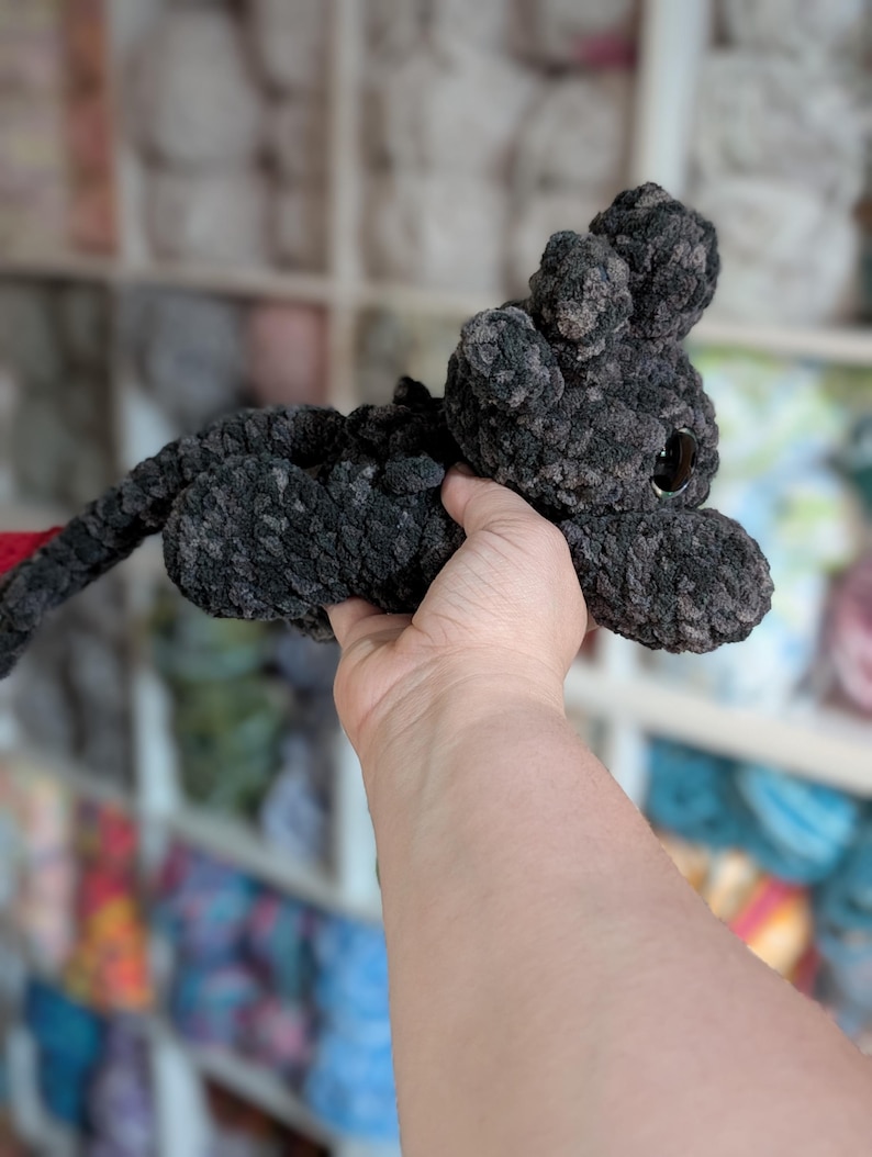 May include: A handmade, dark gray crocheted cat toy with a long tail and large, dark eyes. The cat is made of a soft, textured yarn and has small, rounded ears. The tail is red. The toy is held up in front of a blurred background.