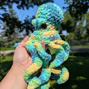 May include: A handmade crochet octopus in shades of blue, green, and yellow. The plush toy has a round head, large black eyes, and eight curly tentacles. The soft, textured yarn creates a whimsical and cuddly appearance, perfect for a child's toy or nursery decor.