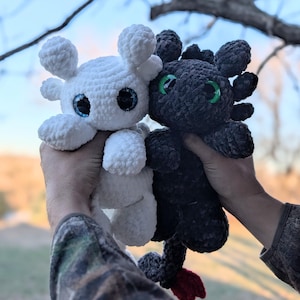 May include: Two crocheted dragon plushies, one white and one black, are held up against a blurred outdoor background. The white dragon has blue eyes, while the black dragon has green eyes. Both have rounded features and are made of a soft, textured yarn.