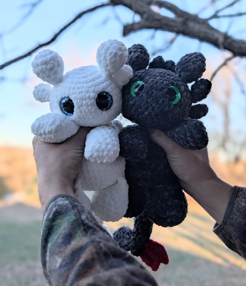 May include: Two crocheted dragon plushies, one white and one black, are held up against a blurred outdoor background. The white dragon has blue eyes, while the black dragon has green eyes. Both have rounded features and are made of a soft, textured yarn.