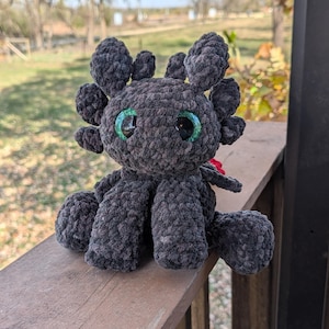 May include: A handmade, dark gray crocheted dragon plushie with large, round, green and black eyes. The plushie has multiple rounded appendages and is sitting on a wooden surface. The dragon has a red accent.