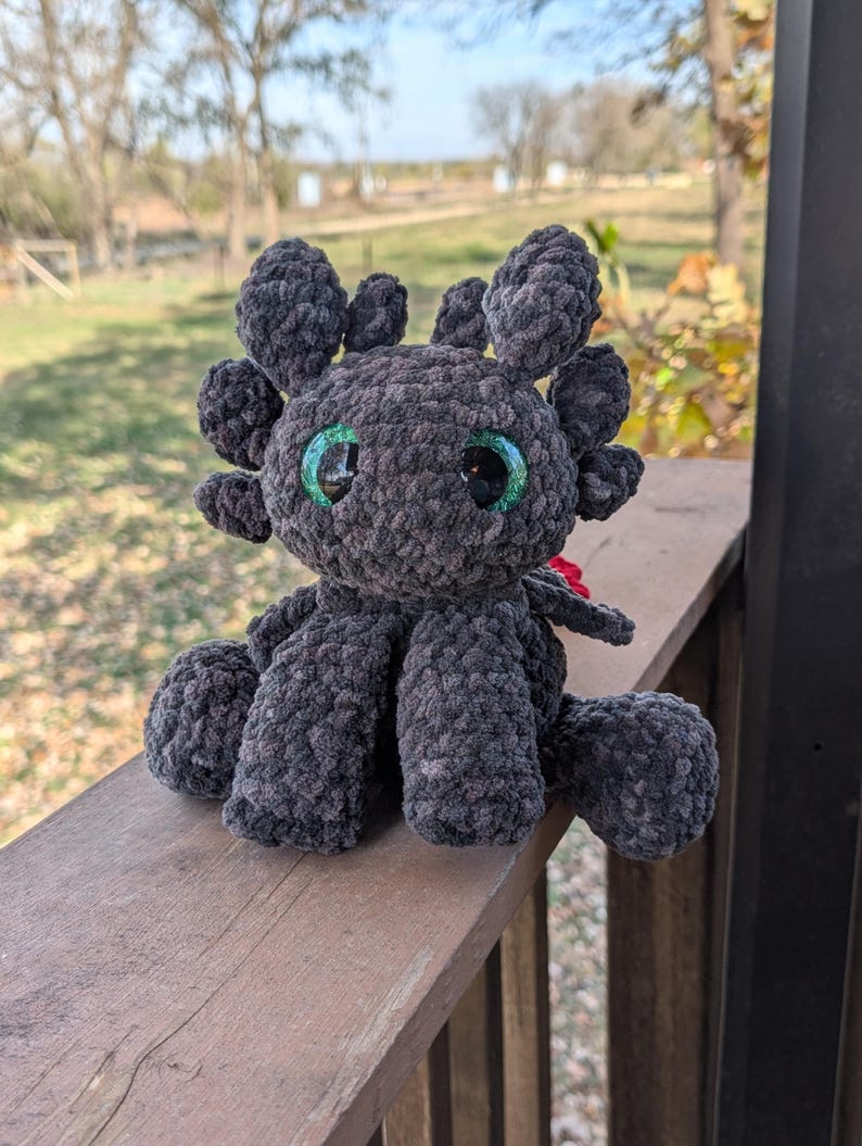 May include: A handmade, dark gray crocheted dragon plushie with large, round, green and black eyes. The plushie has multiple rounded appendages and is sitting on a wooden surface. The dragon has a red accent.
