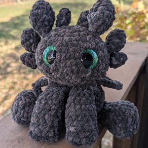 May include: A handmade, dark gray crochet dragon plushie with large, shiny green eyes. The plush features detailed wings, ears, and legs, and is made with a soft, textured yarn. This whimsical dragon is a unique, handcrafted item.