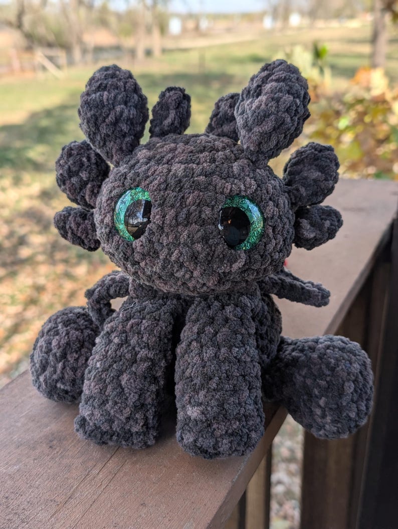 May include: A handmade, dark gray crochet dragon plushie with large, shiny green eyes. The plush features detailed wings, ears, and legs, and is made with a soft, textured yarn. This whimsical dragon is a unique, handcrafted item.