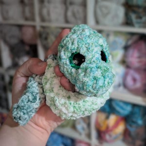 May include: A handmade crochet turtle toy in teal, green, and white. The turtle features large, shiny black eyes with a green border. The soft, textured yarn gives a cuddly appearance, suitable for a child's toy or nursery decoration.