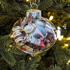 May include: A clear glass Christmas ornament filled with colorful comic book pages. The ornament is hanging on a green Christmas tree.