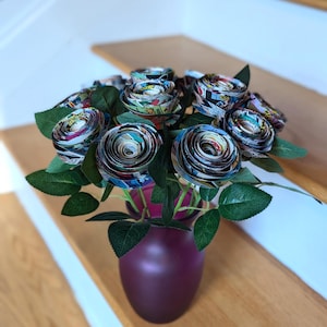May include: A bouquet of comic book paper roses in a deep purple vase. The roses are arranged in a circular pattern and have a variety of colors and patterns.