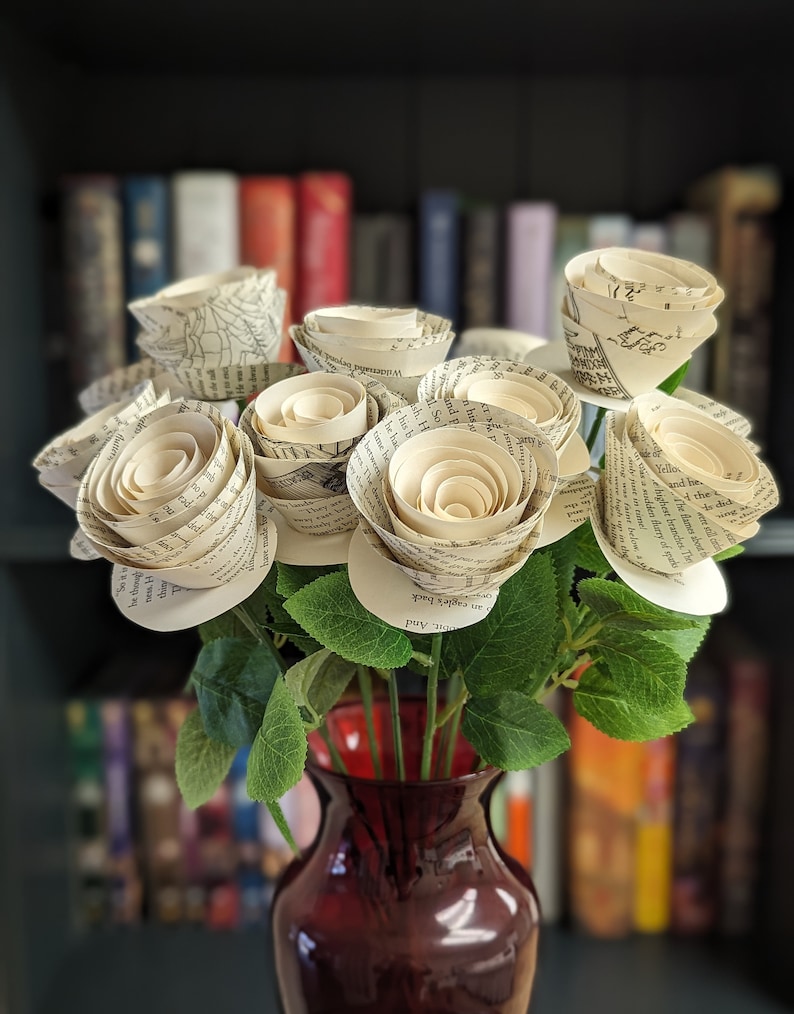A bouquet of roses, in a red glass vase, made from the pages of a book with green stems and leaves in front of an out of focus bookshelf.