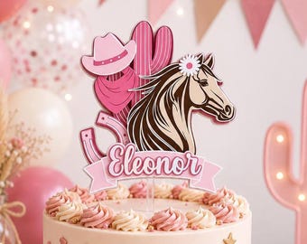 May include: A cake topper featuring a brown horse head, a pink cowboy hat, a pink bandana, and a pink cactus. The design includes a pink banner with the name Eleonor in white script, and a pink horseshoe. The topper is placed on a frosted cake.