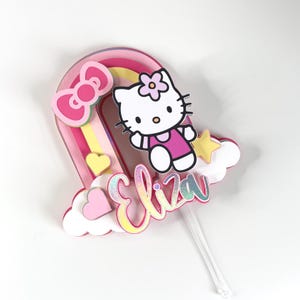Może przedstawiać: Topper na tort w kształcie tęczy w kolorze r&oacute;żowym i ż&oacute;łtym z postacią Hello Kitty, ż&oacute;łtą gwiazdą i imieniem "Eliza" w r&oacute;żowych i tęczowych literach.