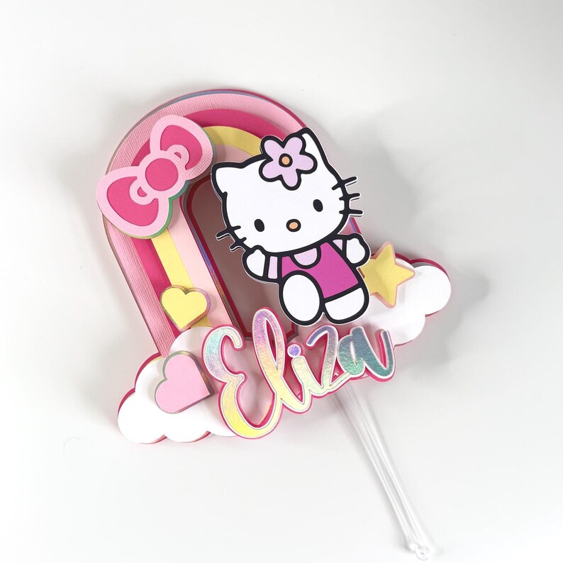 Może przedstawiać: Topper na tort w kształcie tęczy w kolorze r&oacute;żowym i ż&oacute;łtym z postacią Hello Kitty, ż&oacute;łtą gwiazdą i imieniem "Eliza" w r&oacute;żowych i tęczowych literach.