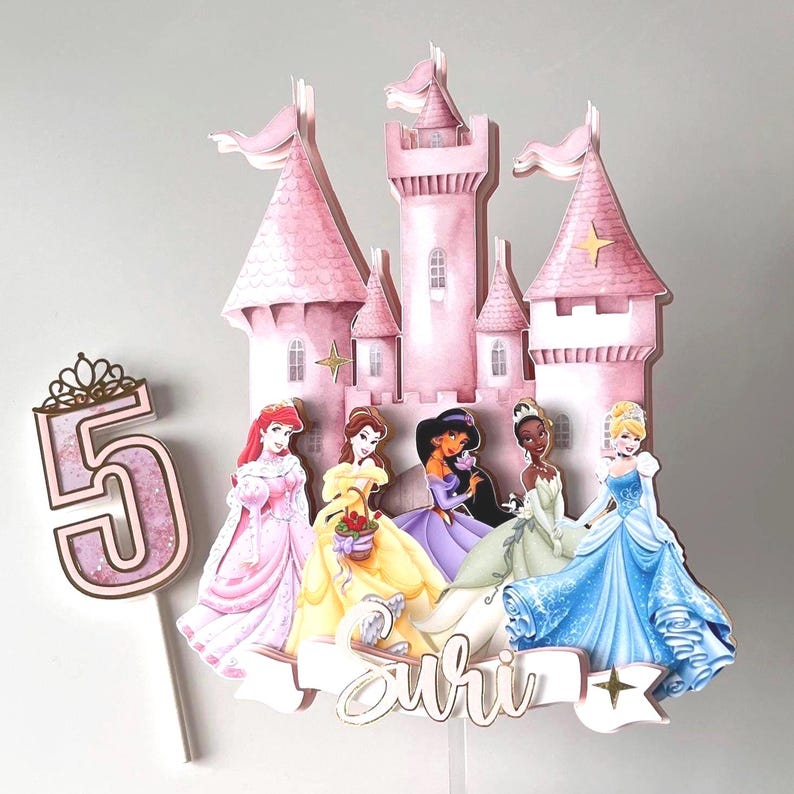 May include: A pink castle cake topper with six Disney princesses, including Ariel, Belle, Jasmine, Tiana, Cinderella, and a pink glitter number 5. The cake topper is decorated with gold stars and the name "Suhi" is written on a banner.