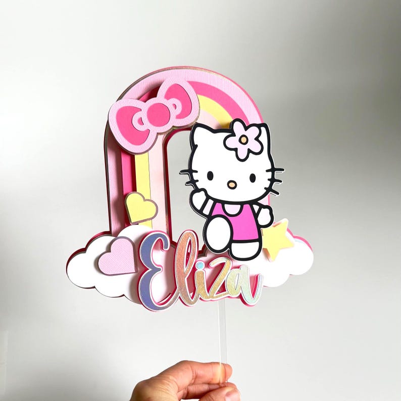 Może przedstawiać: Topper na tort w kształcie tęczy w kolorze r&oacute;żowym i ż&oacute;łtym z postacią Hello Kitty, r&oacute;żową kokardką i imieniem "Eliza" w błyszczących literach.