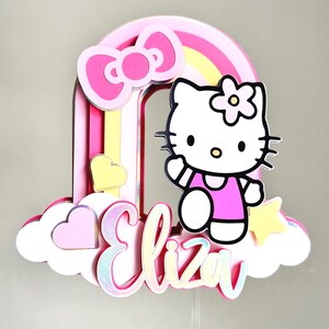 Może przedstawiać: Topper na tort w kształcie tęczy w kolorze r&oacute;żowym i ż&oacute;łtym z postacią Hello Kitty, r&oacute;żową kokardką i imieniem "Eliza" w r&oacute;żowych literach z brokatem.