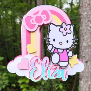 Może przedstawiać: Topper na tort w kształcie tęczy w kolorze r&oacute;żowym i ż&oacute;łtym z postacią Hello Kitty, r&oacute;żową kokardką i imieniem "Eliza" w błyszczącym foncie.