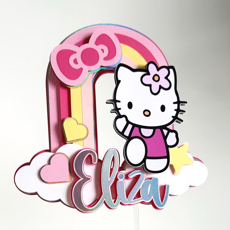 Może przedstawiać: Topper na tort w kształcie tęczy w kolorze r&oacute;żowym, ż&oacute;łtym i niebieskim z postacią Hello Kitty w kolorze białym, r&oacute;żową kokardką i ż&oacute;łtą gwiazdą. Imię "Eliza" jest napisane na białej chmurze błyszczącymi literami.