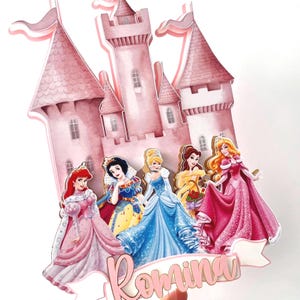 May include: A pink castle cake topper with five Disney princesses: Ariel, Snow White, Cinderella, Belle, and Aurora. The cake topper is decorated with gold accents and the name "Romina" is written in pink cursive.