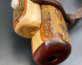Könnte beinhalten: Eine handgefertigte Holzpfeife mit natürlicher Holzmaserung. Die Pfeife hat ein dunkelbraunes Mundstück, einen hellen Holzkörper und einen Lederriemen. Ein kleiner, weißer Stein ist in das Holz eingelassen.
