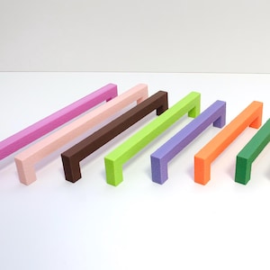 Può includere: Una collezione di maniglie colorate per armadi in varie tonalità, tra cui rosa, marrone, verde, viola, arancione, giallo, grigio e blu. Ogni maniglia ha una barra rettangolare con un'impugnatura ad angolo retto, perfetta per mobili.