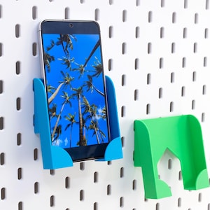 以下が含まれることがあります： スマートフォンは、白いペグボードに取り付けられた明るい青色のホルダーに固定されています。携帯電話には熱帯の風景が表示されます。別の緑色のホルダーも表示されます。ホルダーは、アイテムを整理して表示するように設計されています。