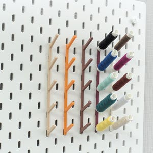 Puede incluir: Un tablero de clavijas blanco con clavijas de madera y carretes de hilo de varios colores, incluyendo rojo, azul, verde, amarillo y marrón.