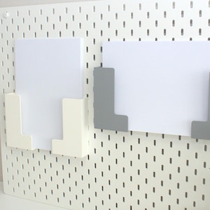 Può includere: Due organizzatori di carta bianchi con cornici in plastica grigia e bianca montati su una parete di bacheca bianca.